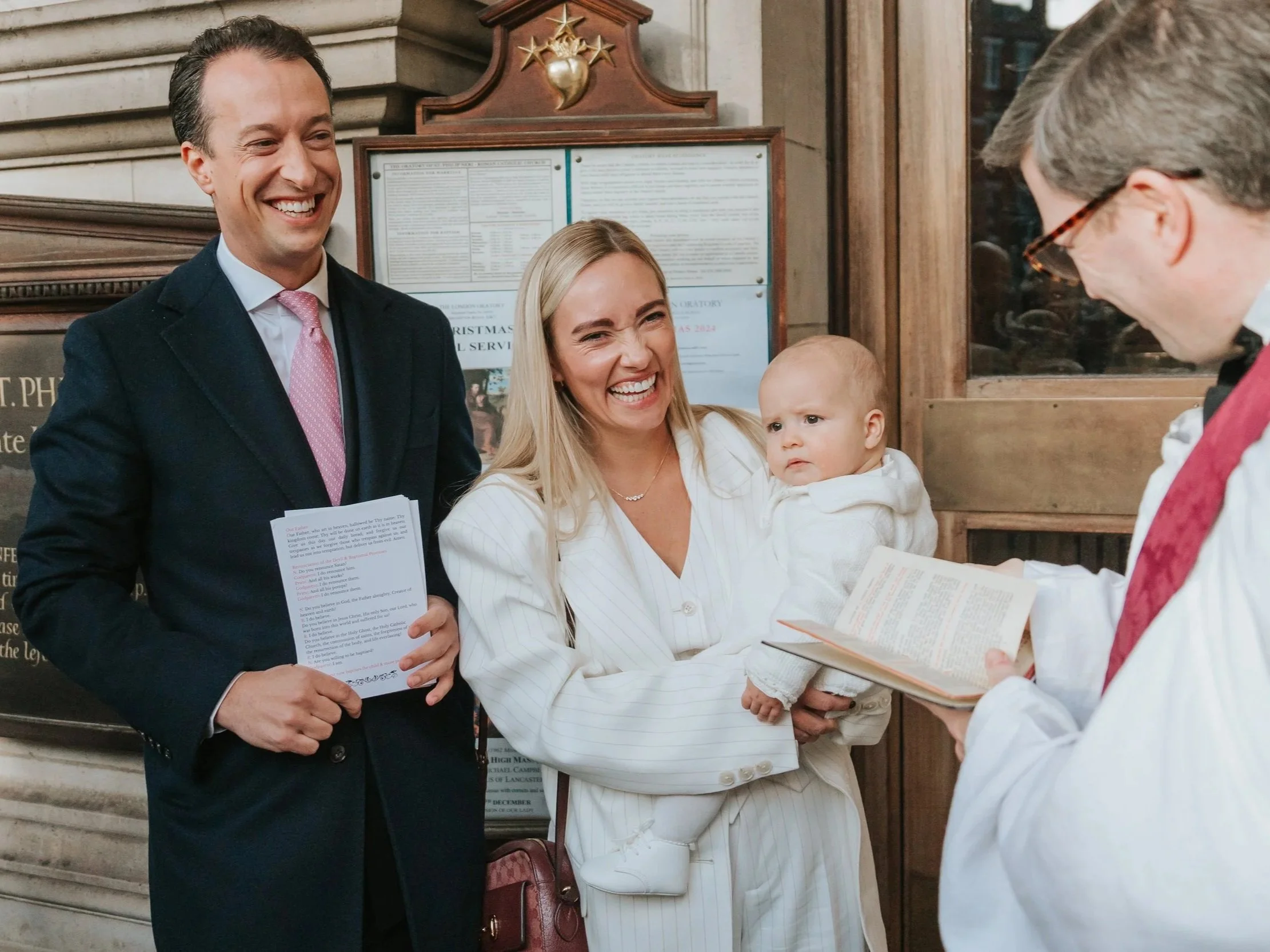  Smiling parents holding their baby in a christening gown inside Brompton Oratory, London 