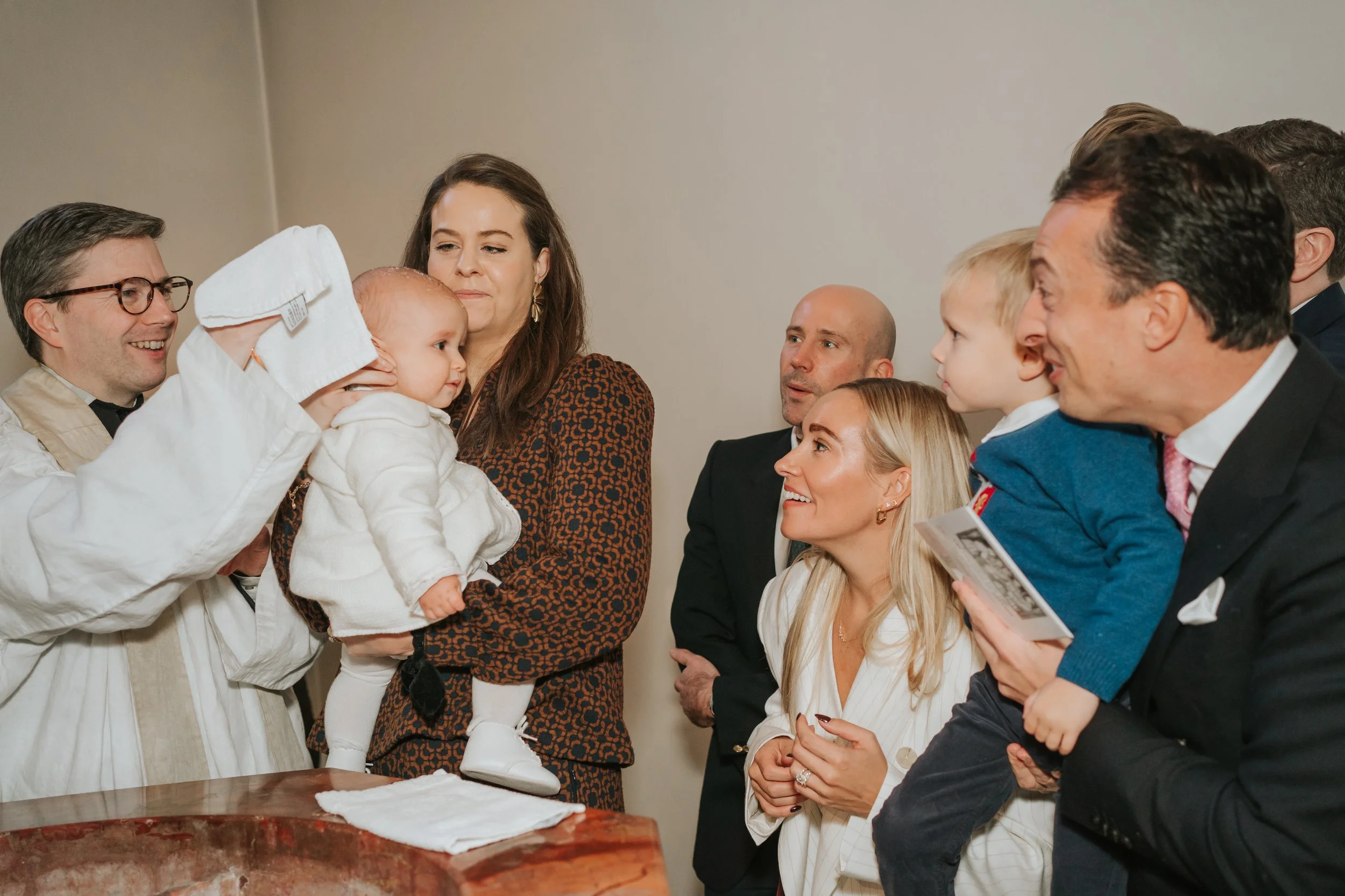  Priest pouring holy water over a baby’s head during a christening at Brompton Oratory, with family and godparents gathered closely around the baptismal font. 
