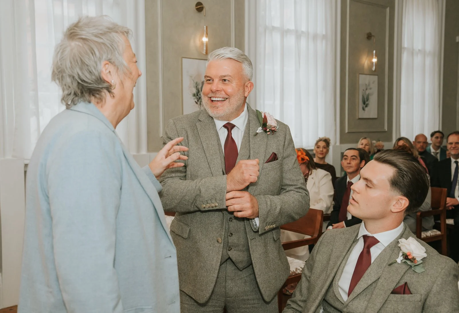  The groom shares a laugh with guests before the ceremony at Chelsea Old Town Hall, dressed in a grey tweed suit with burgundy tie and boutonniere. 