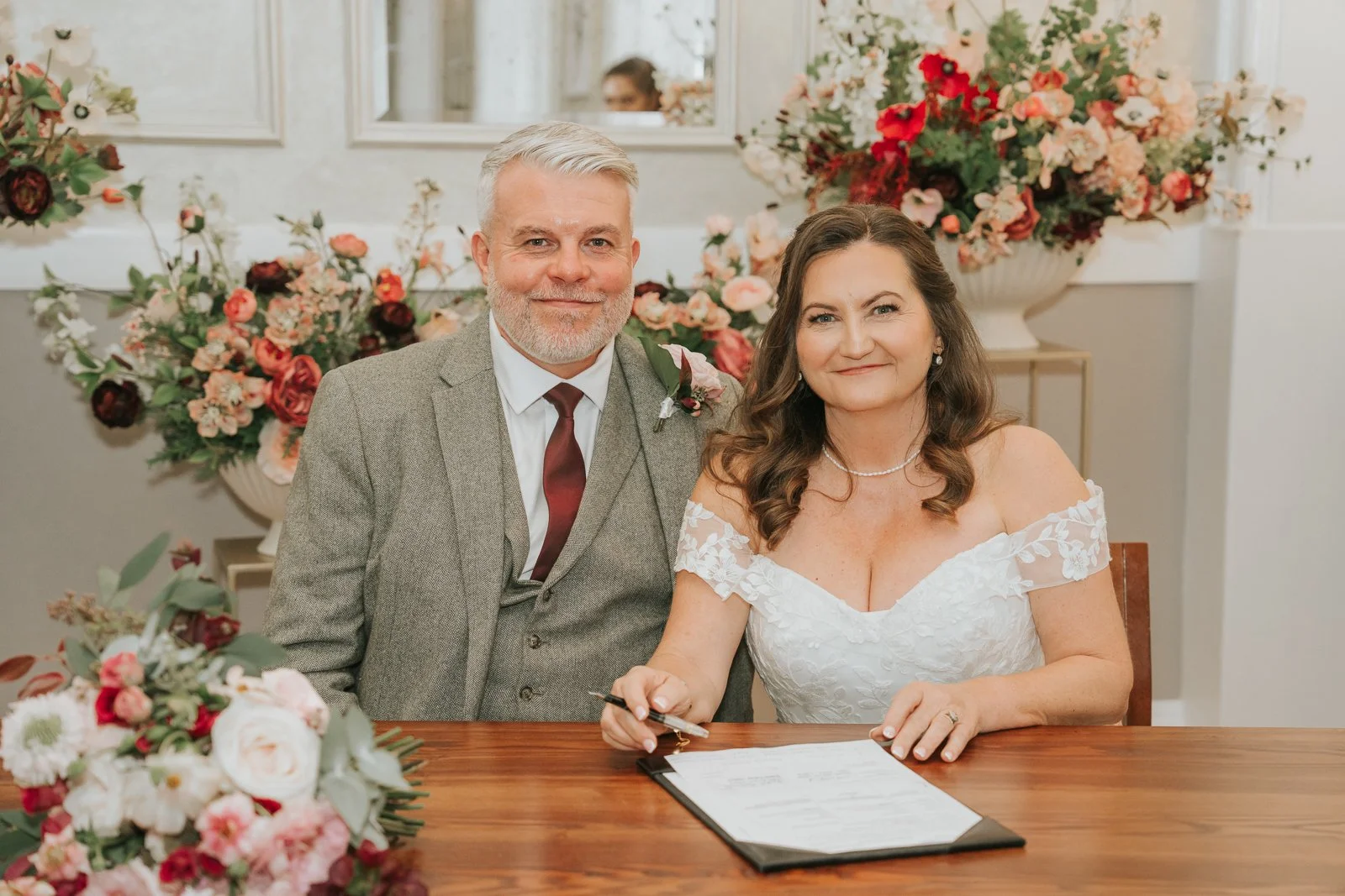  Bride and groom signing the marriage register at Chelsea Old Town Hall, surrounded by elegant floral arrangements in soft pink, cream, and red tones. 