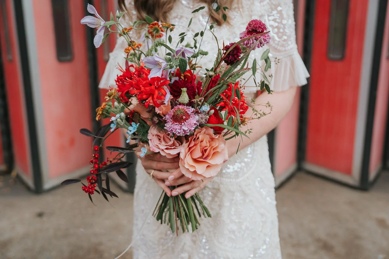  Close-up of the bride’s bouquet featuring red dahlias, blush roses, and pink scabiosa against the textured beaded fabric of her wedding dress. 