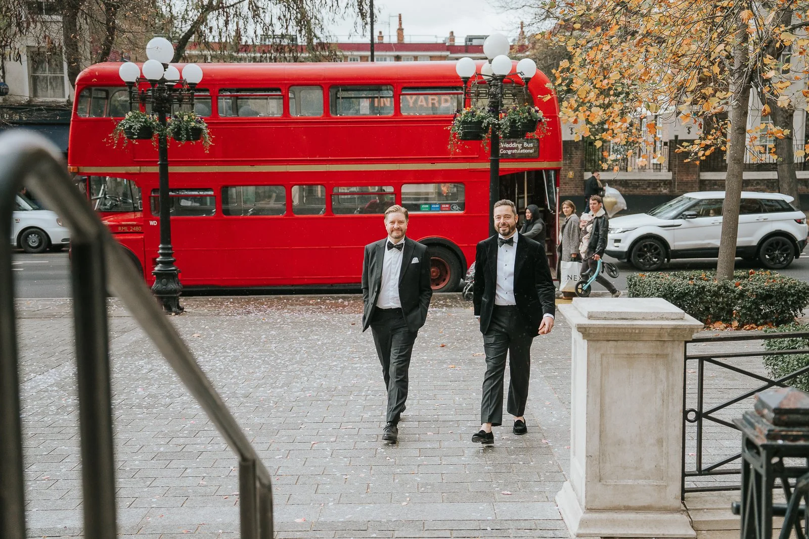  Groom and groomsman in black ties walking towards Islington Town Hall with a classic red London bus behind them. 