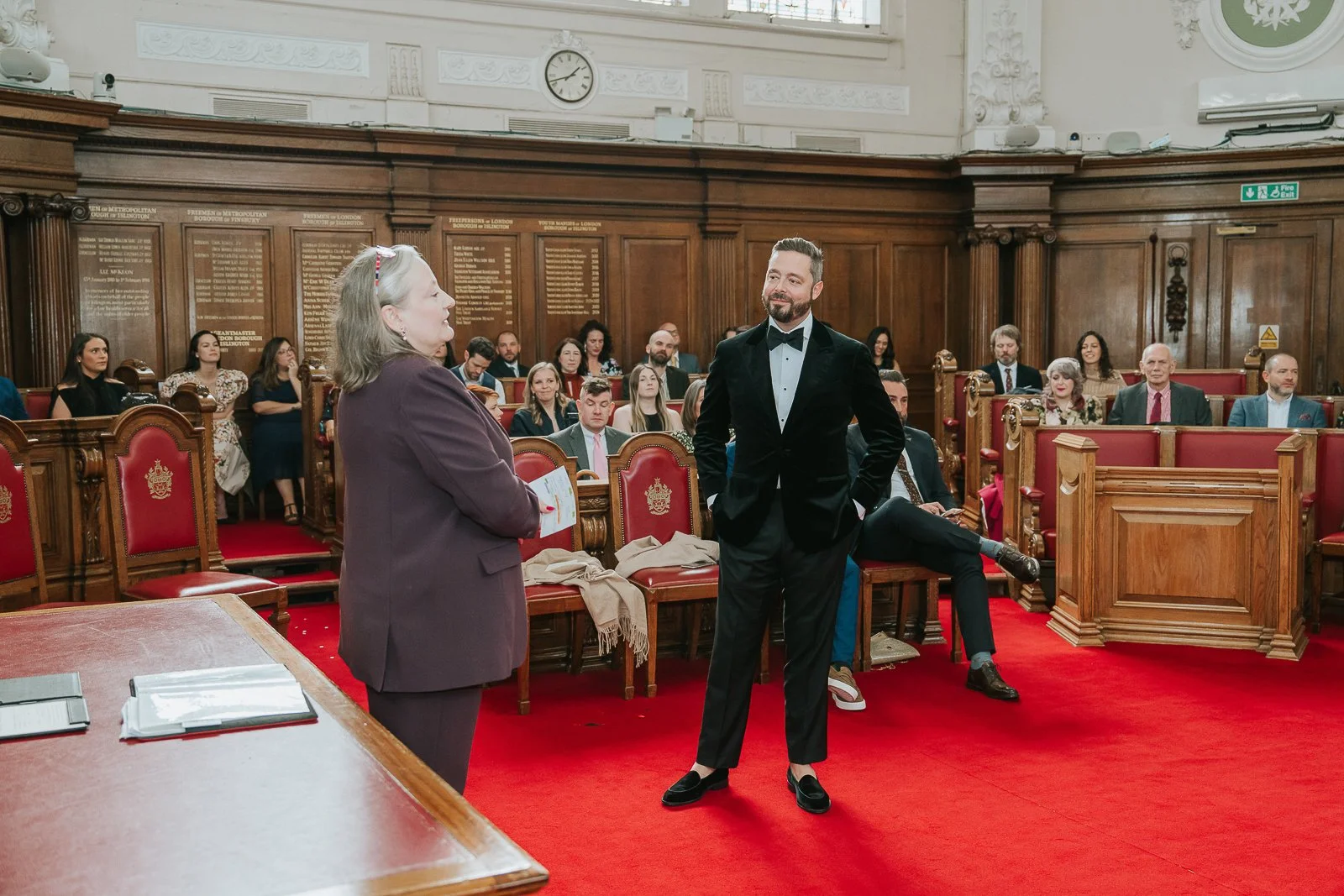  Groom smiling as the registrar begins the ceremony inside the Council Chamber at Islington Town Hall, surrounded by guests in the historic wood-panelled room. 