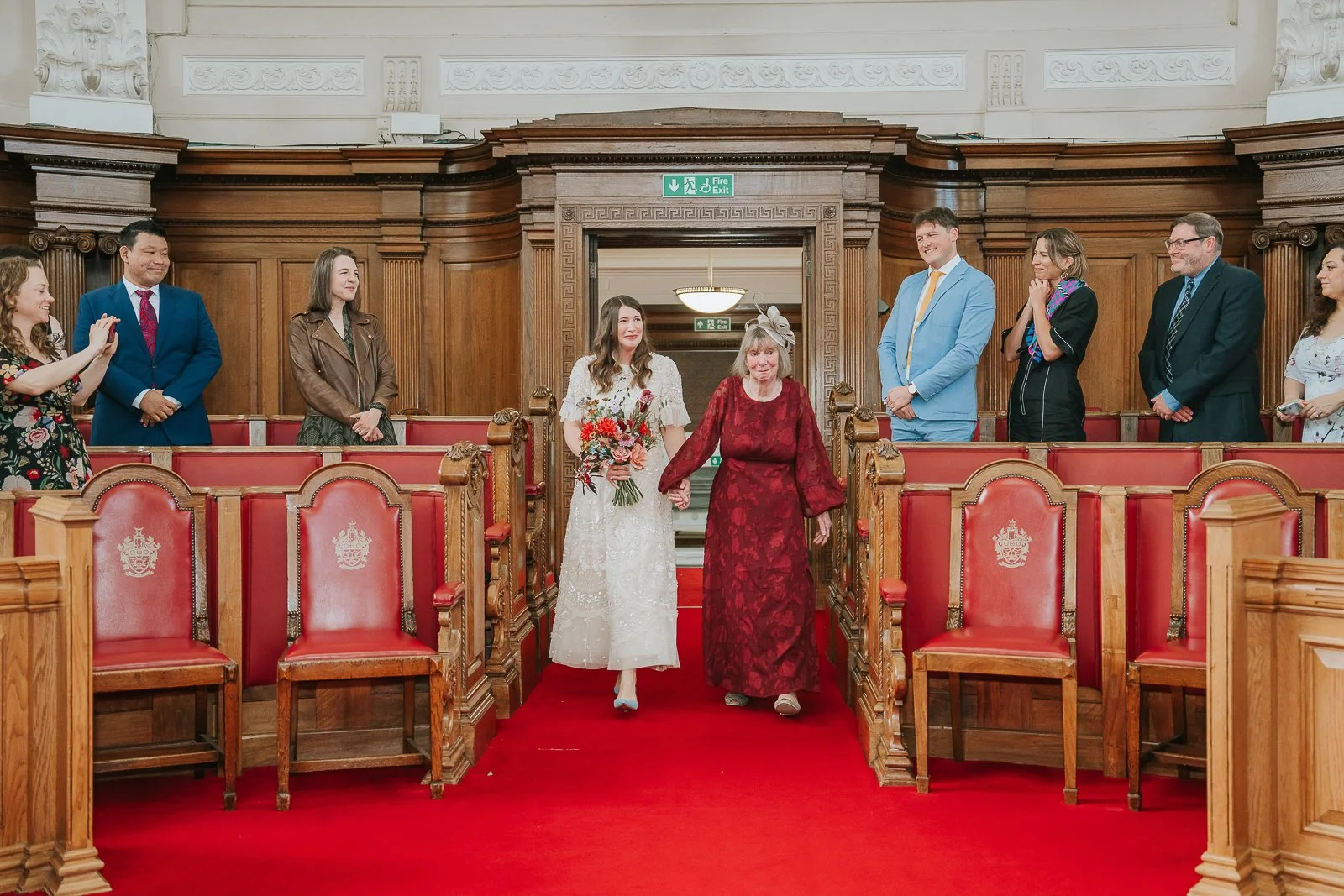  Bride walking down the red carpet aisle at Islington Town Hall with her mother, holding a colourful bouquet of autumn flowers. 