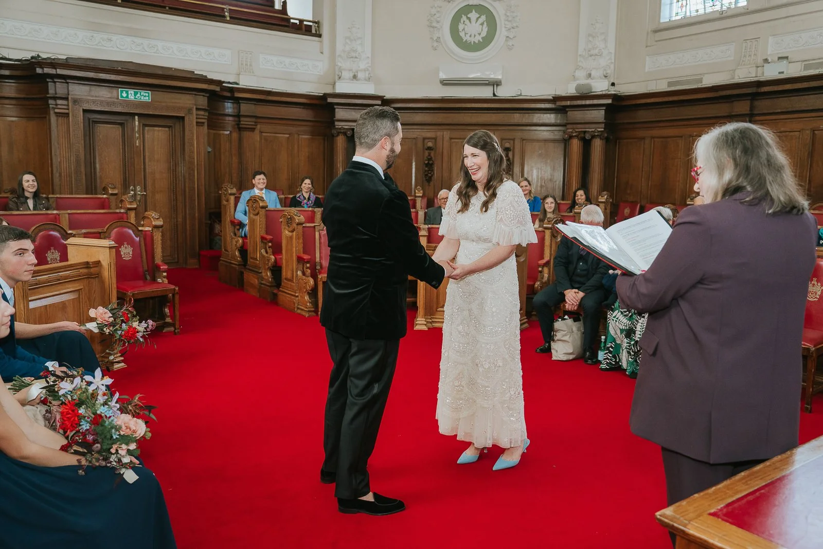 Bride and groom exchanging vows and holding hands in the Council Chamber at Islington Town Hall, surrounded by friends and family. 
