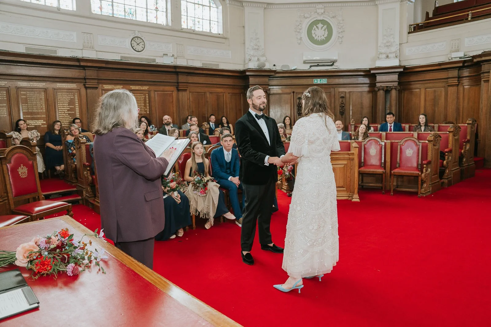  Bride and groom exchanging vows and holding hands in the Council Chamber at Islington Town Hall, surrounded by friends and family. 