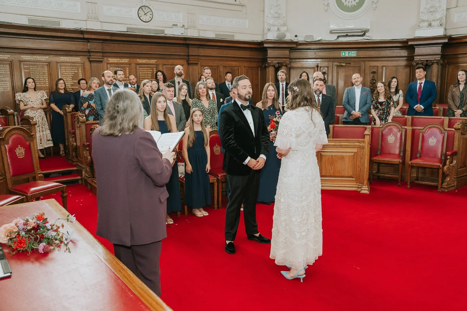  Bride and groom exchanging vows and holding hands in the Council Chamber at Islington Town Hall, surrounded by friends and family. 