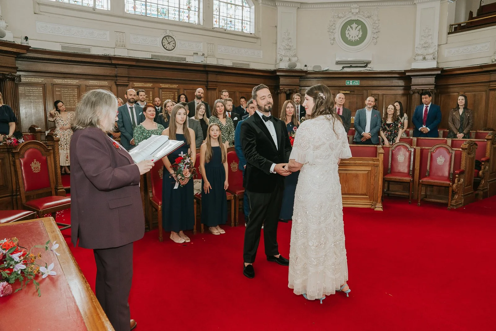  Bride and groom exchanging vows and holding hands in the Council Chamber at Islington Town Hall, surrounded by friends and family. 