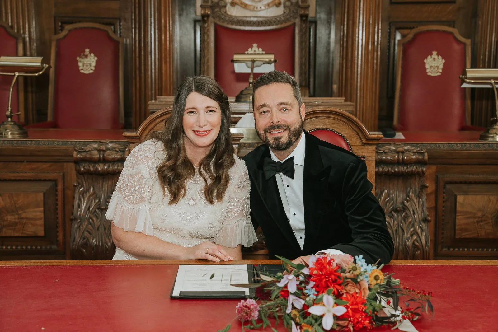  Couple smiling as they sign the marriage register at Islington Town Hall, with bright florals and red leather chairs in the background. 