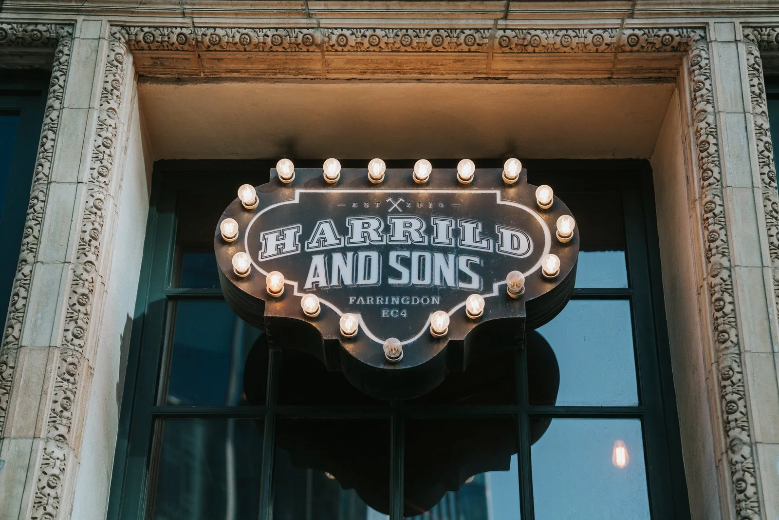  The illuminated Harrild &amp; Sons exterior sign glowing above the entrance, framed by ornate stone detailing and large windows. 