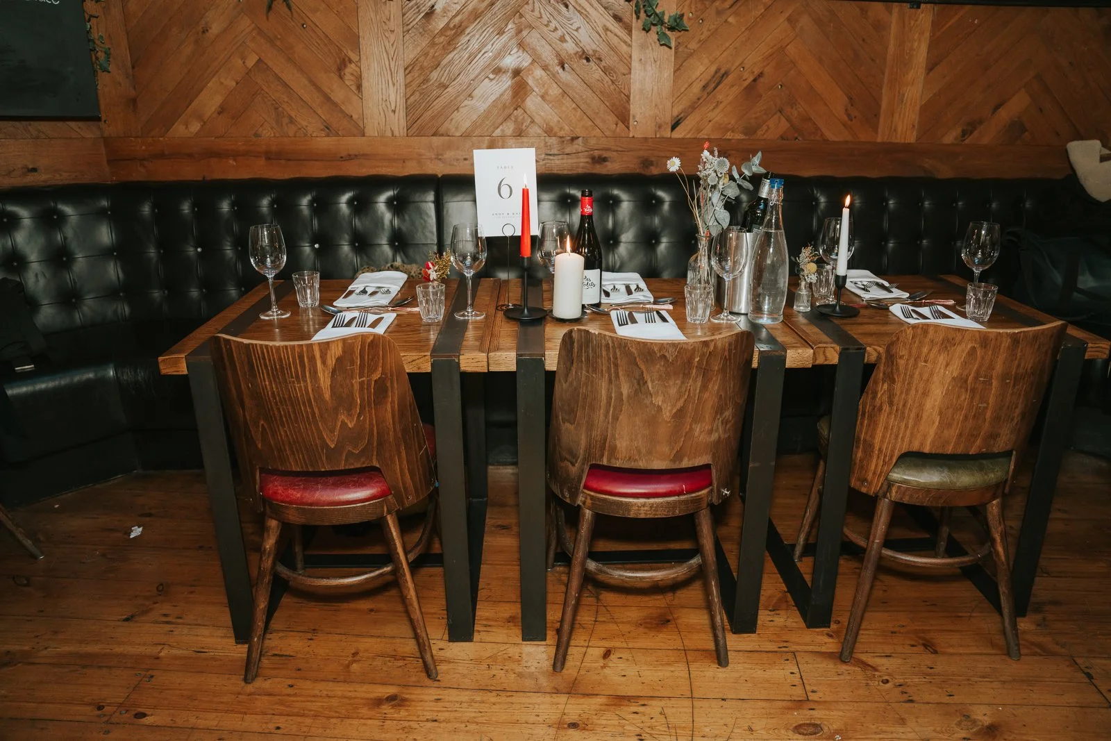  A long wooden dining table set for a wedding meal at Harrild &amp; Sons, with candles, florals and rustic chairs against an exposed brick wall and leather banquette. 
