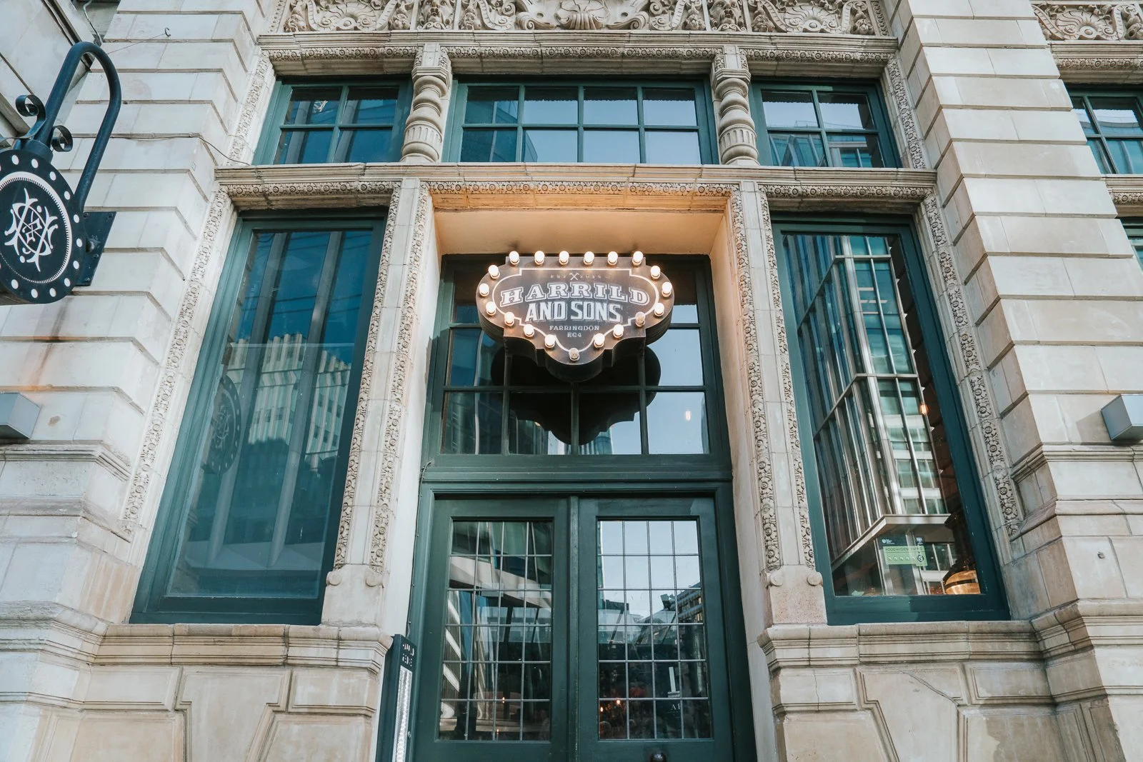  The illuminated Harrild &amp; Sons exterior sign glowing above the entrance, framed by ornate stone detailing and large windows. 