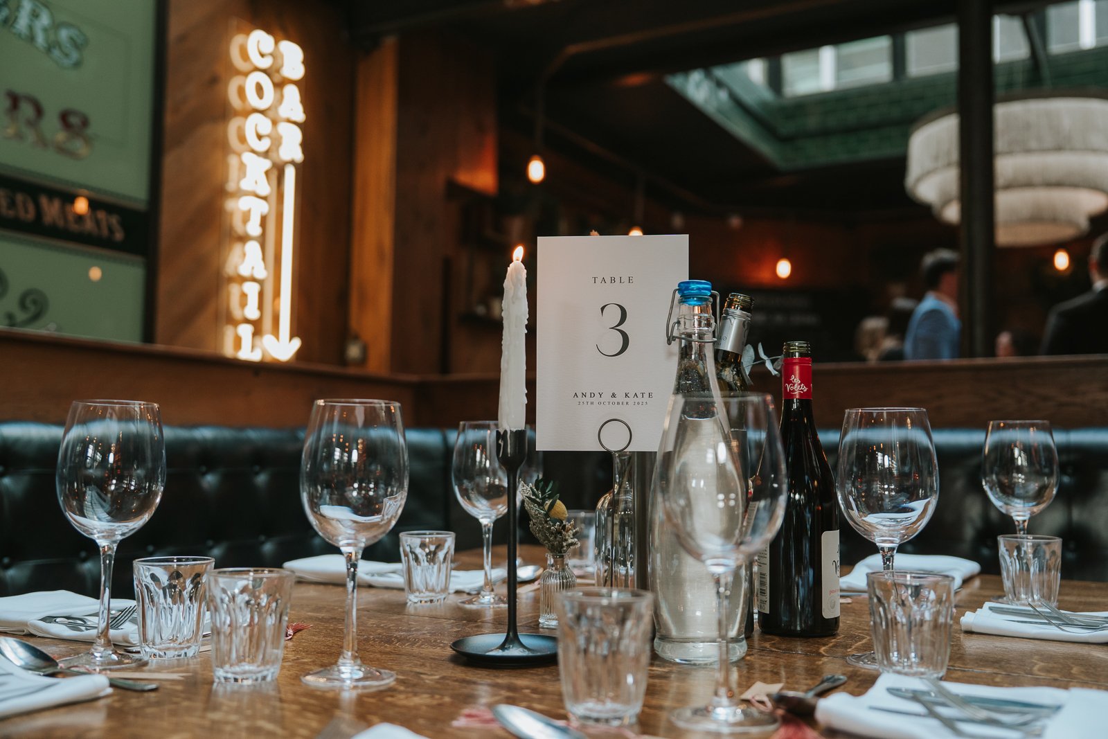  A neatly laid Harrild &amp; Sons wedding dining table with a “Table 1” sign, candles, glassware and bottled drinks, with the cocktail bar neon sign in the background. 