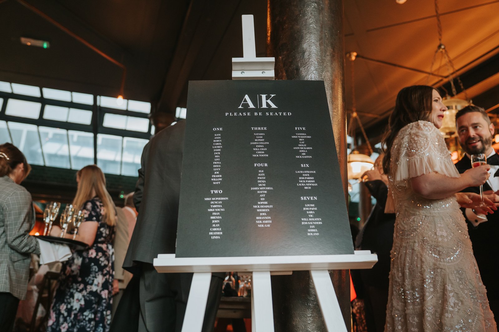  A modern black seating plan displayed on an easel inside Harrild &amp; Sons as guests in formal outfits chat and enjoy drinks around it. 