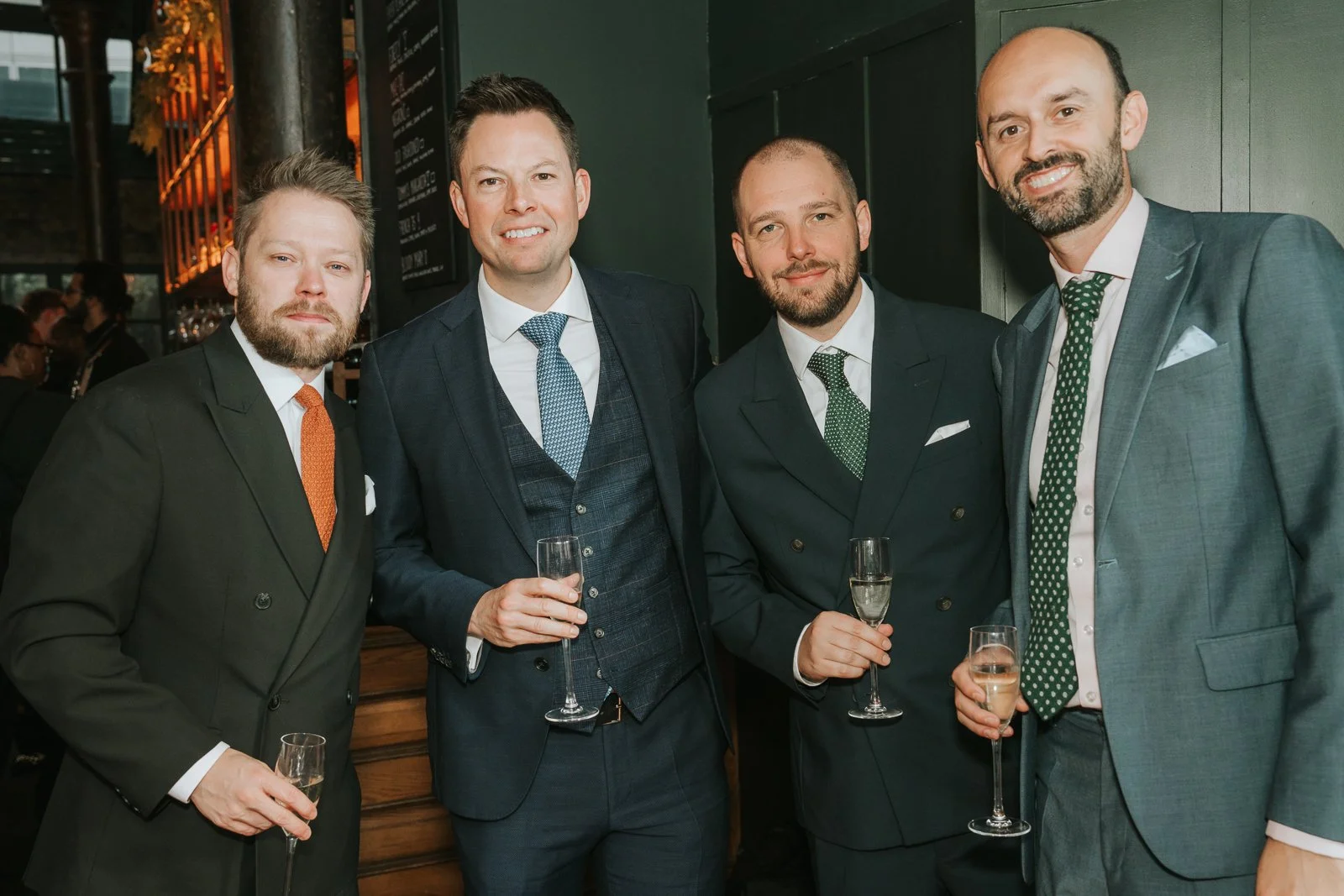  Four wedding guests in suits standing together with champagne inside Harrild &amp; Sons, smiling in front of the dark green bar area and warm ambient lights. 