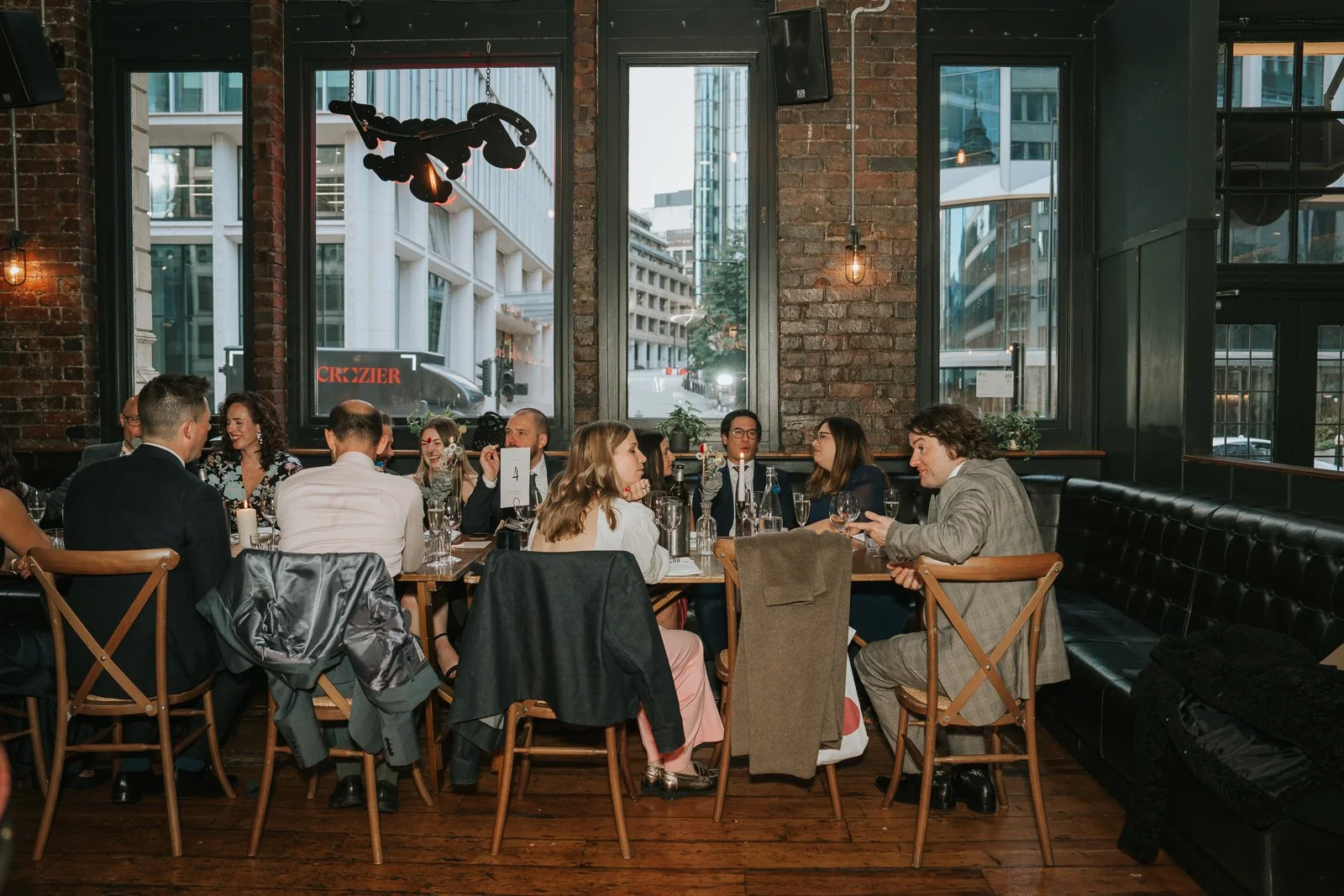  Wedding guests seated at a long dining table inside Harrild &amp; Sons, chatting over candlelight with large industrial windows showing the city street outside. 