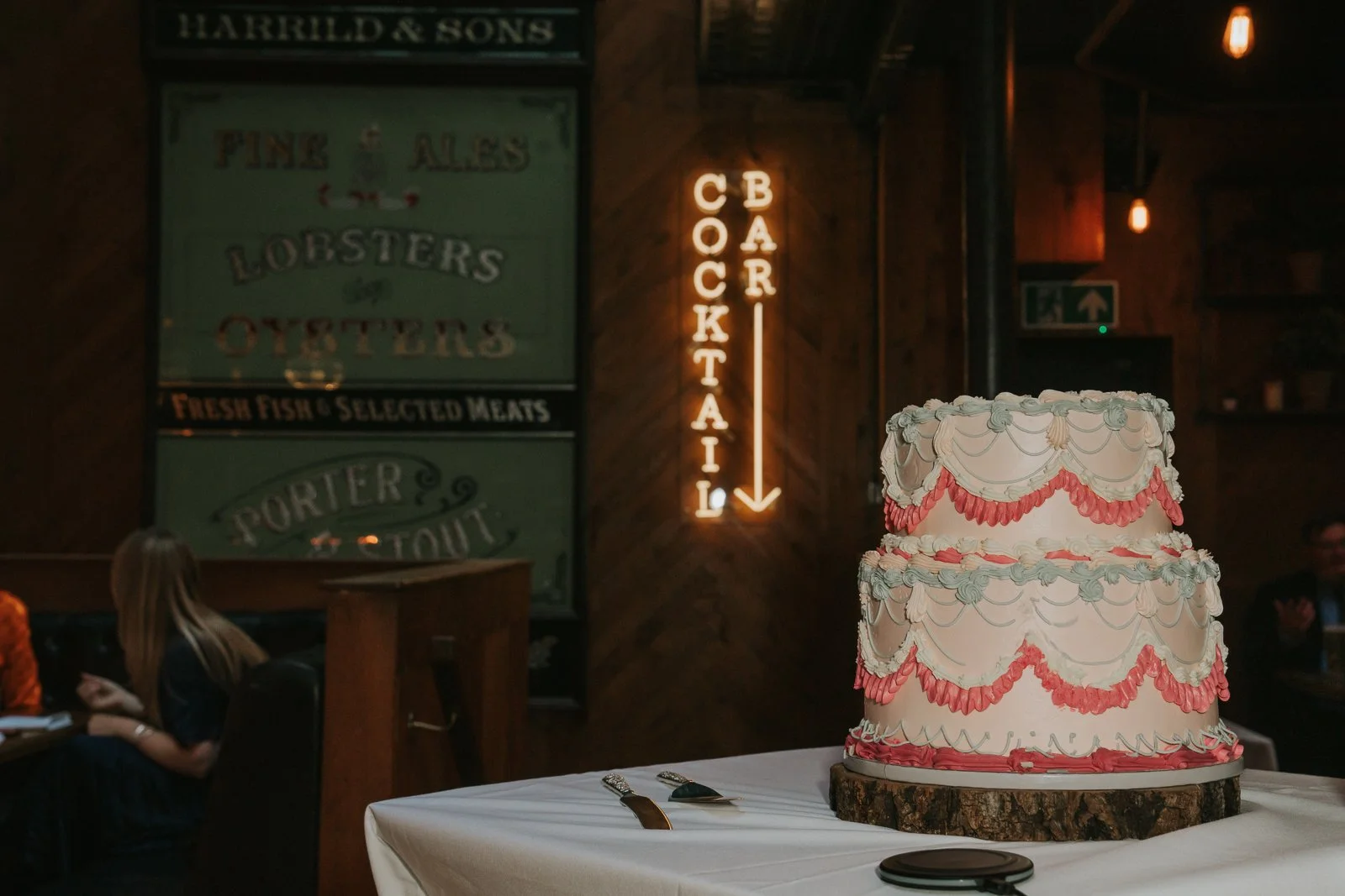 A two-tier vintage-style wedding cake displayed on a wooden board inside Harrild &amp; Sons, with the illuminated “Cocktail Bar” sign glowing behind it. 