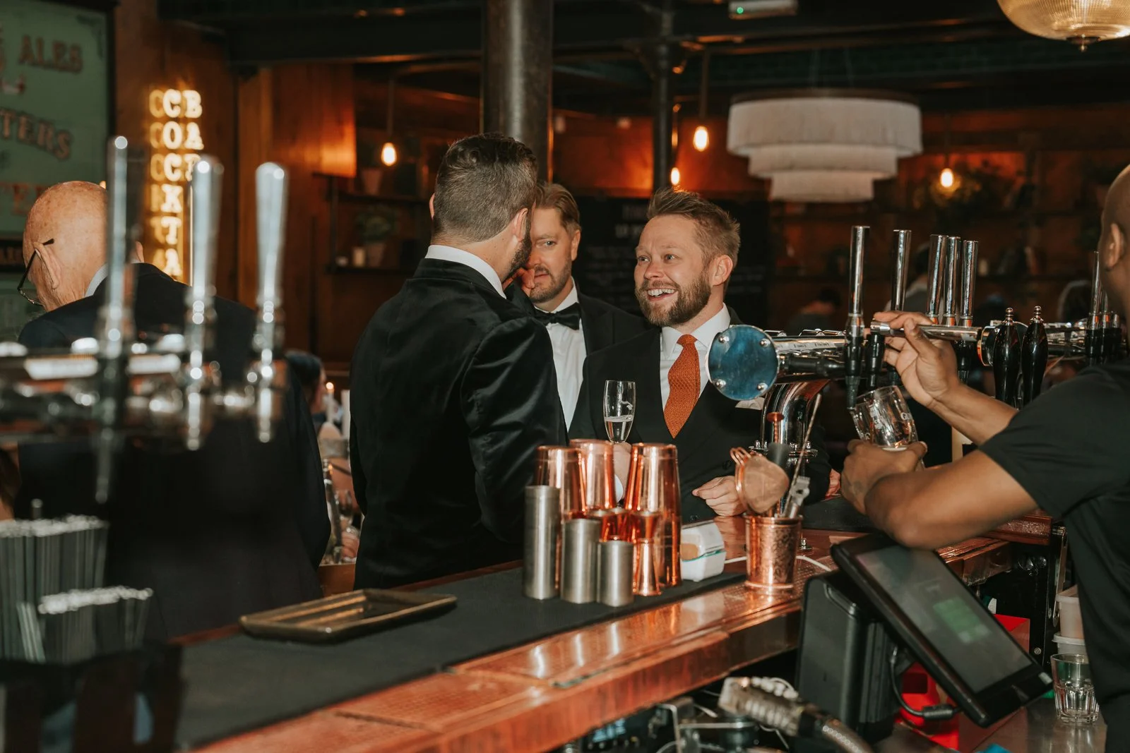  Wedding guests chatting and laughing at the bar inside Harrild &amp; Sons, with copper barware, beer taps and warm lighting creating a lively evening atmosphere. 