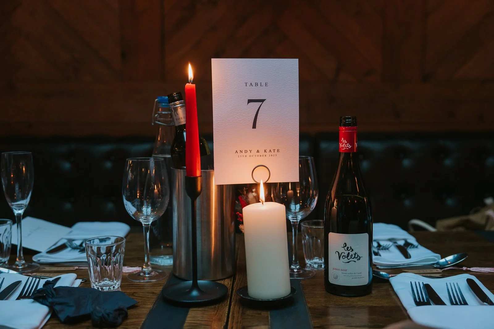  A candlelit wedding table at Harrild &amp; Sons featuring a “Table 7” sign, wine bottles, glassware and a glowing pillar candle against the dark, cosy booth seating. 
