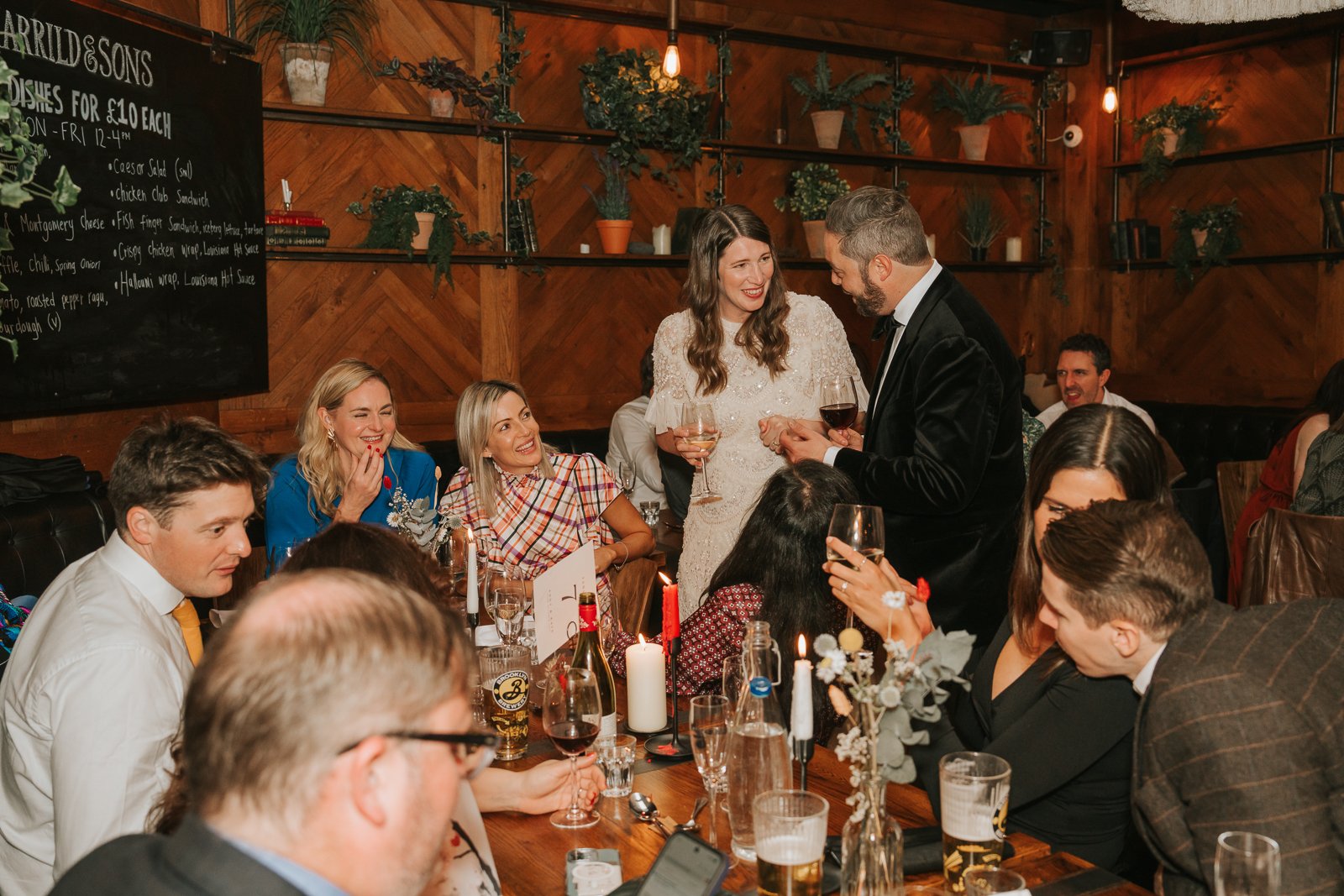  Candid moment of the couple chatting with guests at a long table inside Harrild &amp; Sons, surrounded by friends smiling and laughing in the cosy wood-panelled dining area. 