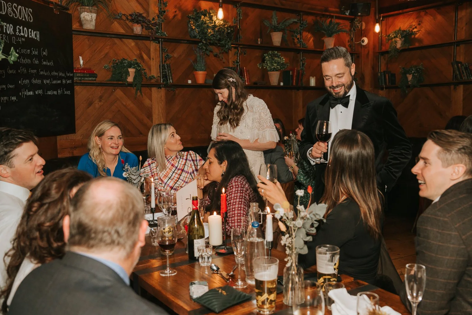  The couple mingling with guests around a candlelit dinner table at Harrild &amp; Sons, with glasses of wine, rustic décor and warm ambient lighting filling the scene. 