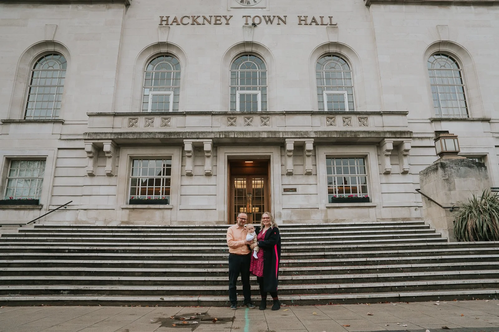  Family portrait on the steps of Hackney Town Hall, with parents holding their baby beneath the iconic façade after the naming ceremony. 