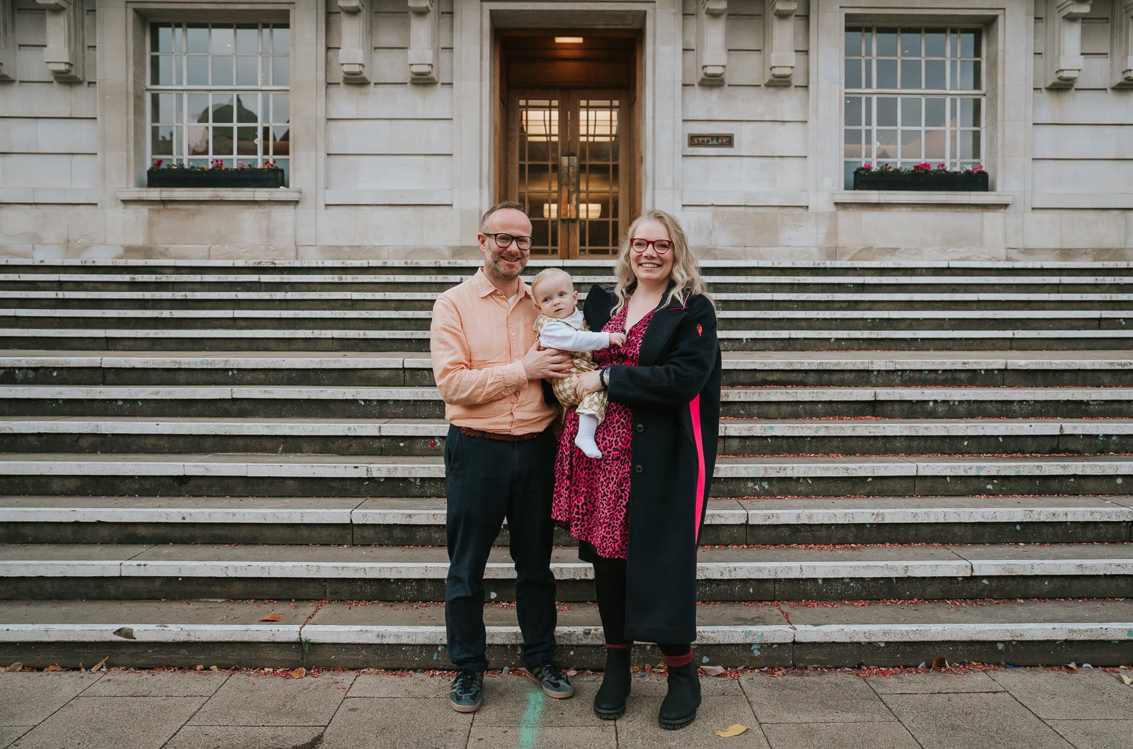  Close-up family photo on the Hackney Town Hall steps, with parents smiling and holding their baby in front of the building’s grand entrance. 