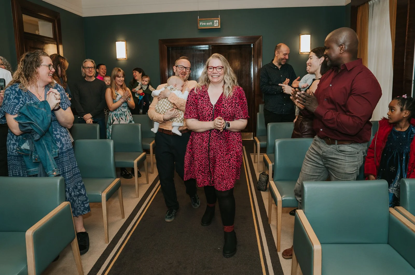  Parents walking down the aisle at Hackney Town Hall with their baby as guests clap and smile at the start of the naming ceremony. 
