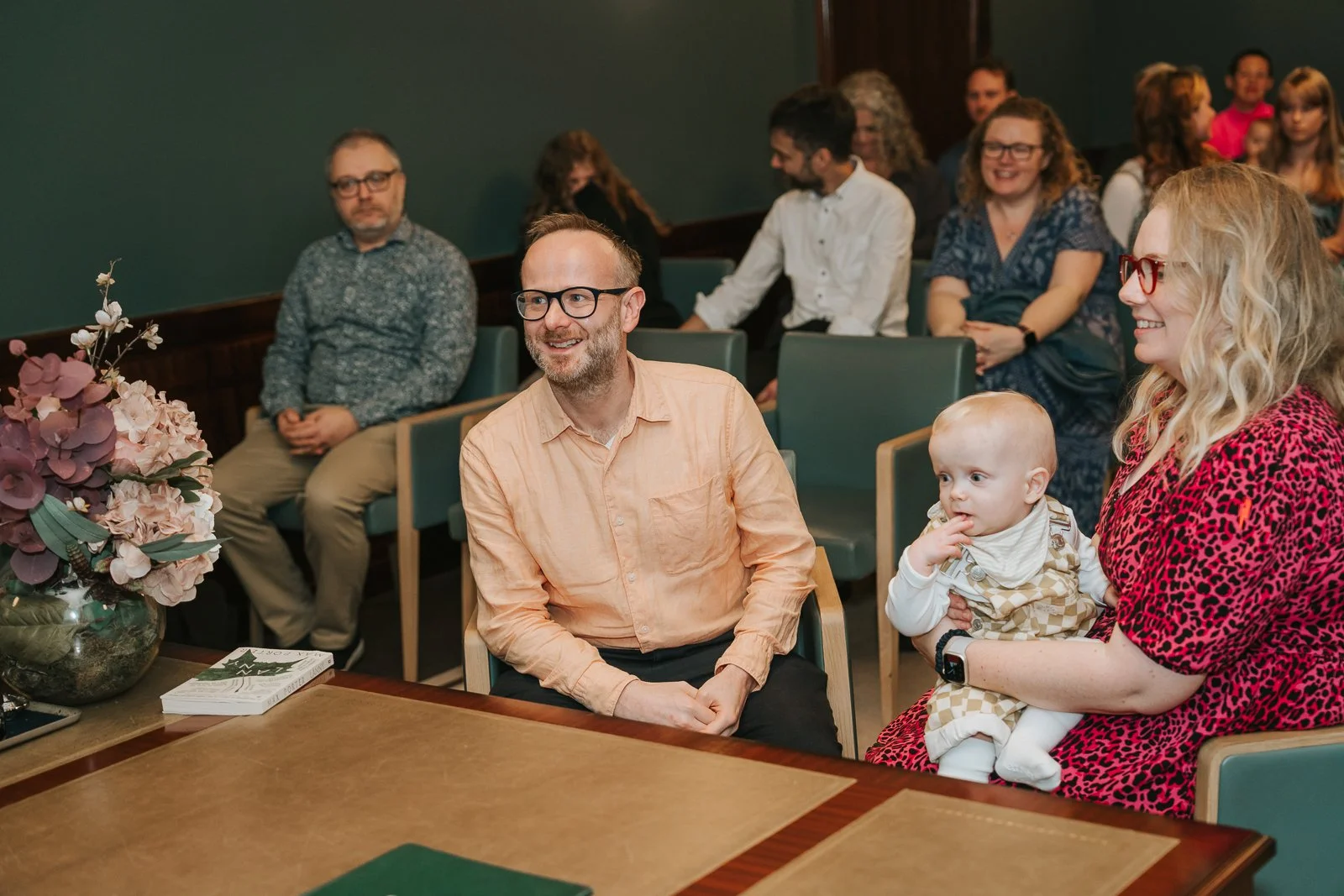  Parents sitting together at the naming ceremony desk in Hackney Town Hall with their baby on their lap, surrounded by smiling guests. 