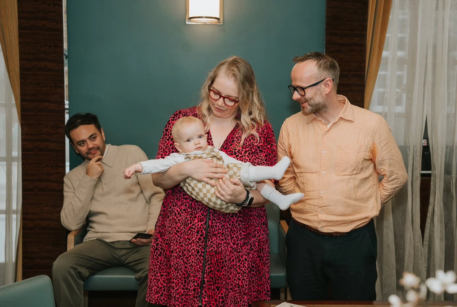  Parents standing together during the ceremony as one holds their baby across their arms in the bright Hackney Town Hall room. 