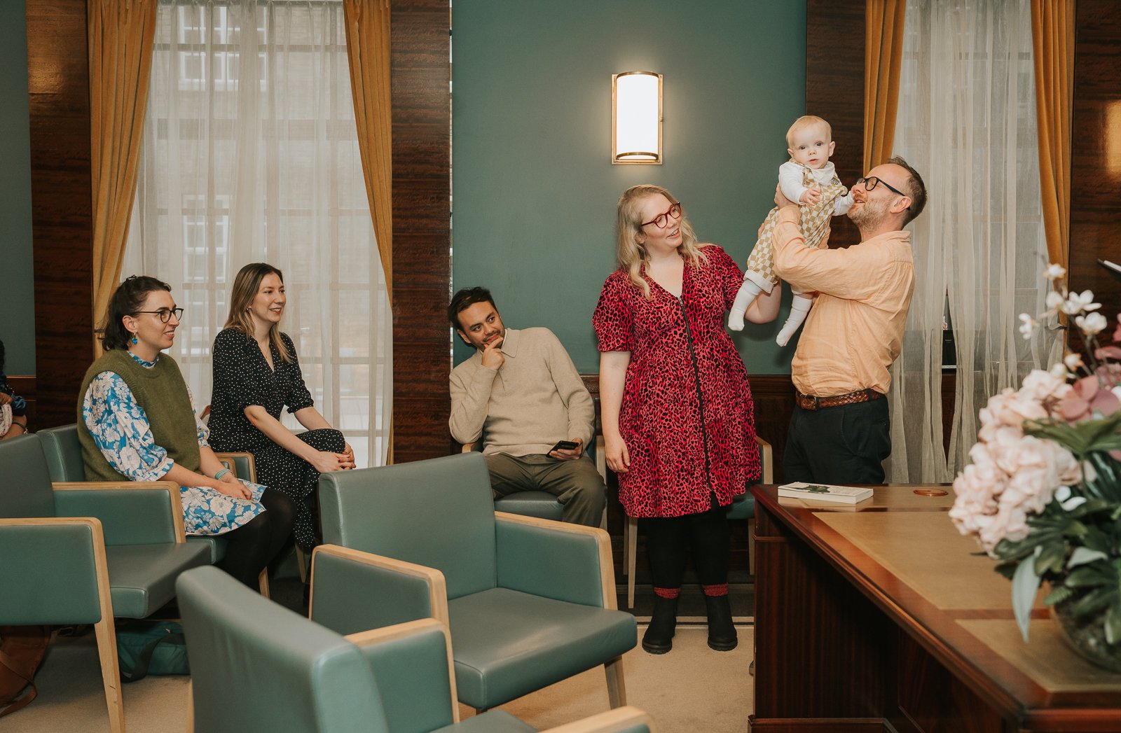  Parents and guests smiling as a parent lifts their baby up during a joyful moment at the Hackney Town Hall naming ceremony. 