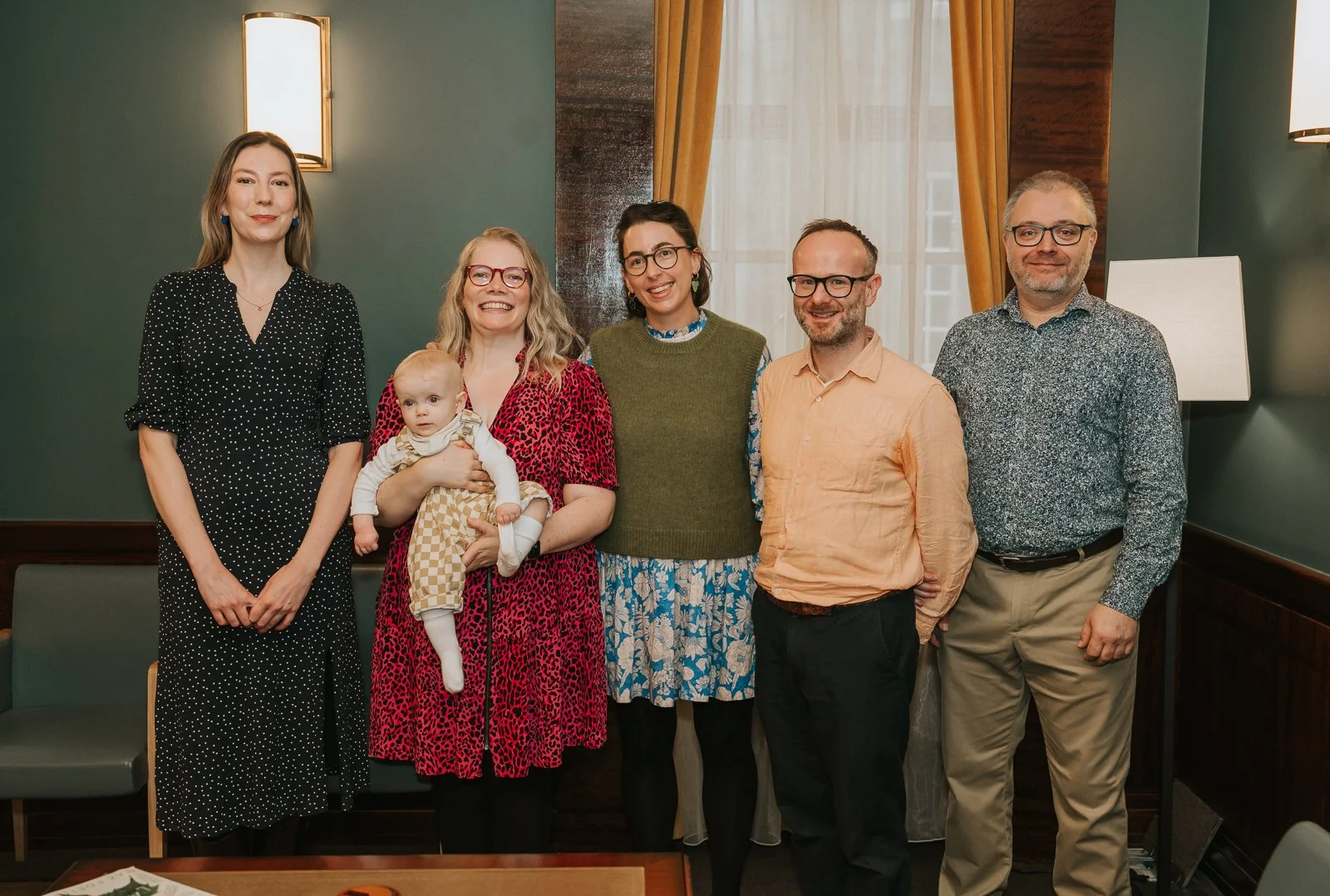  Group portrait of the baby, parents, and the child’s guideparents standing together at the front of the Hackney Town Hall ceremony room. 