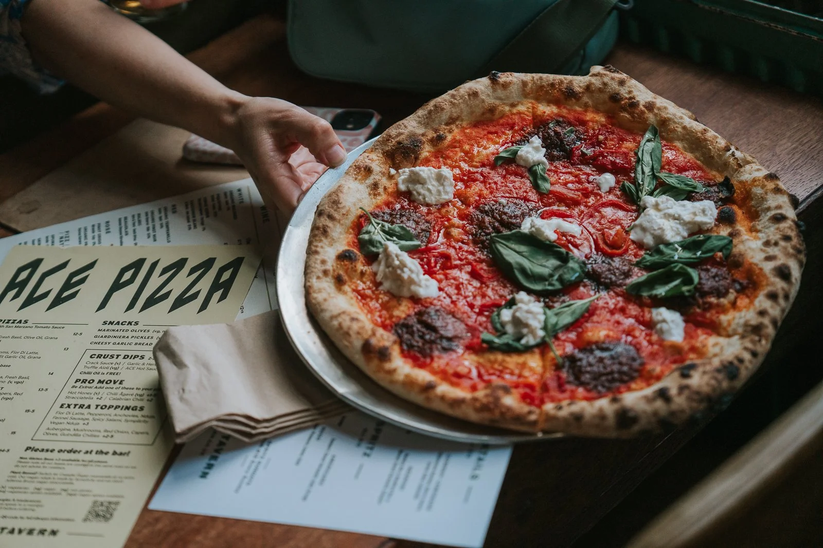  A hand placing down a pizza topped with basil and dollops of cheese, with Ace Pizza menus scattered on the table. 