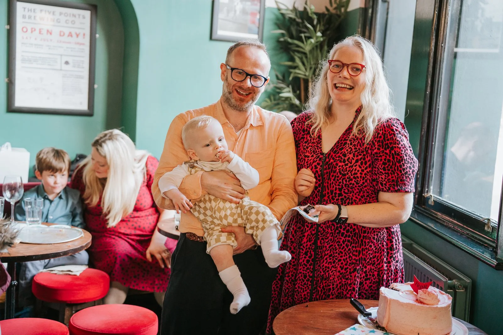  Parents smiling with their baby inside The Pembury Tavern, standing beside the pink celebration cake as guests relax in the background. 