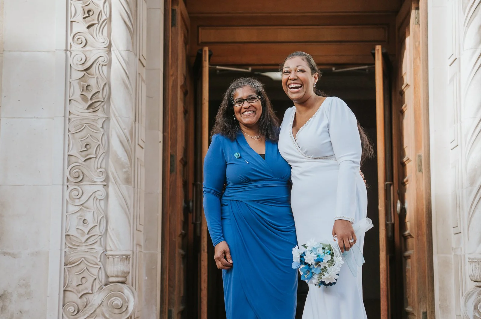  Bride sharing a joyful hug with mum outside Wandsworth Town Hall, both smiling widely against the carved stone doorway. 