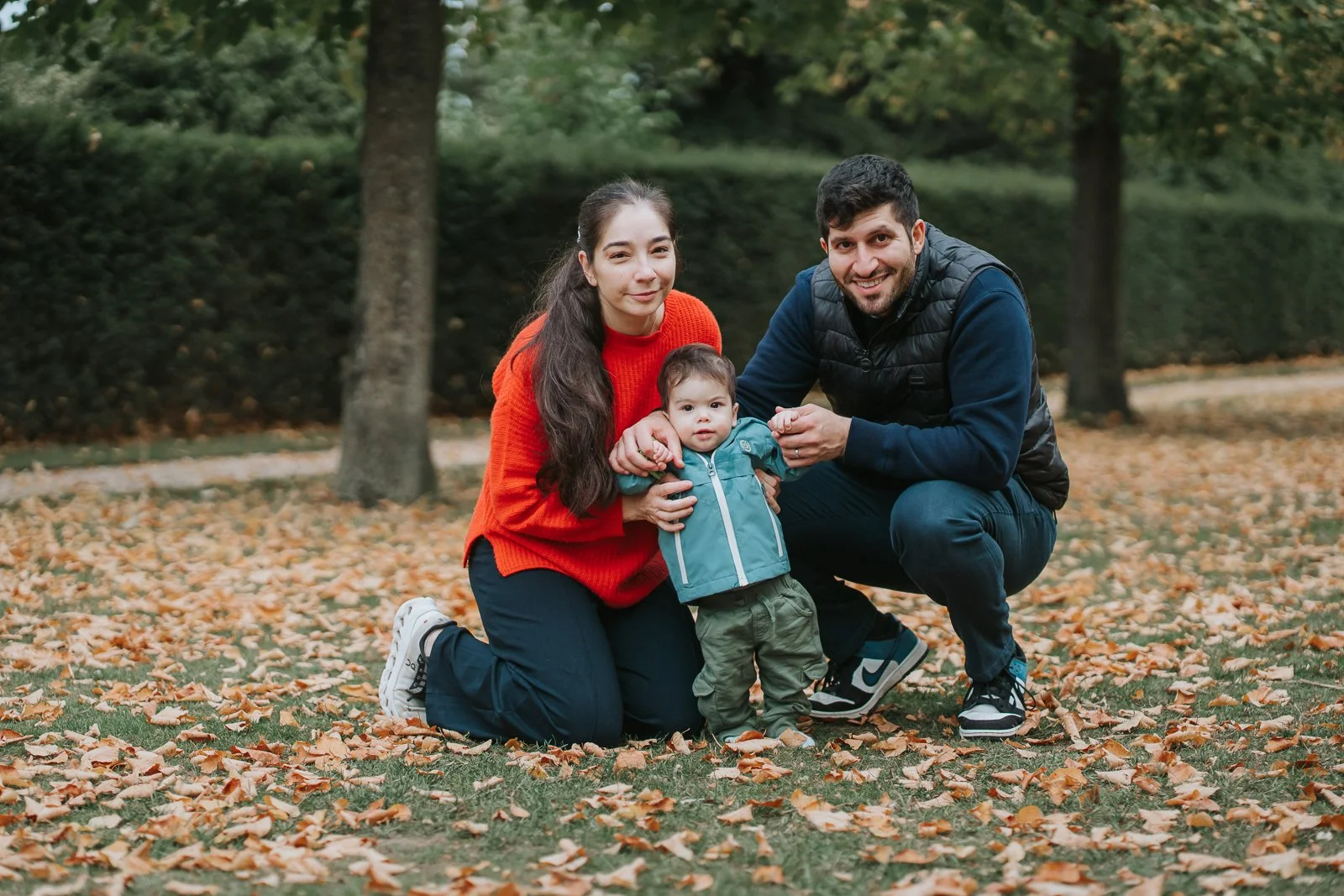  A family kneeling together among autumn leaves at Chiswick House &amp; Gardens, helping their toddler stand during a relaxed West London family photoshoot. 