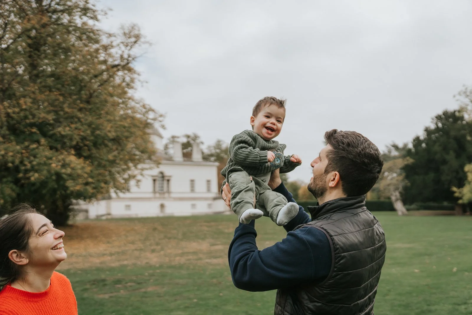  A dad lifting his smiling baby in front of the white Chiswick House villa, with the mum laughing nearby during a joyful West London family photoshoot. 