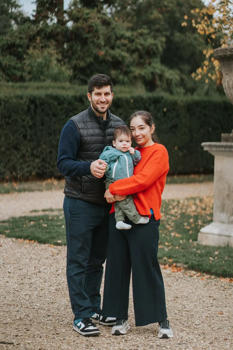  A family standing together on the gravel avenue at Chiswick House &amp; Gardens, holding their baby and smiling during their West London family photoshoot. 