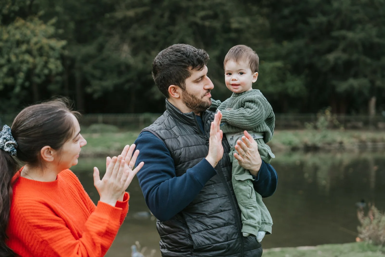  A dad holding his toddler by the lake at Chiswick House &amp; Gardens while the mum claps in the background, captured during a playful family photography session in West London. 