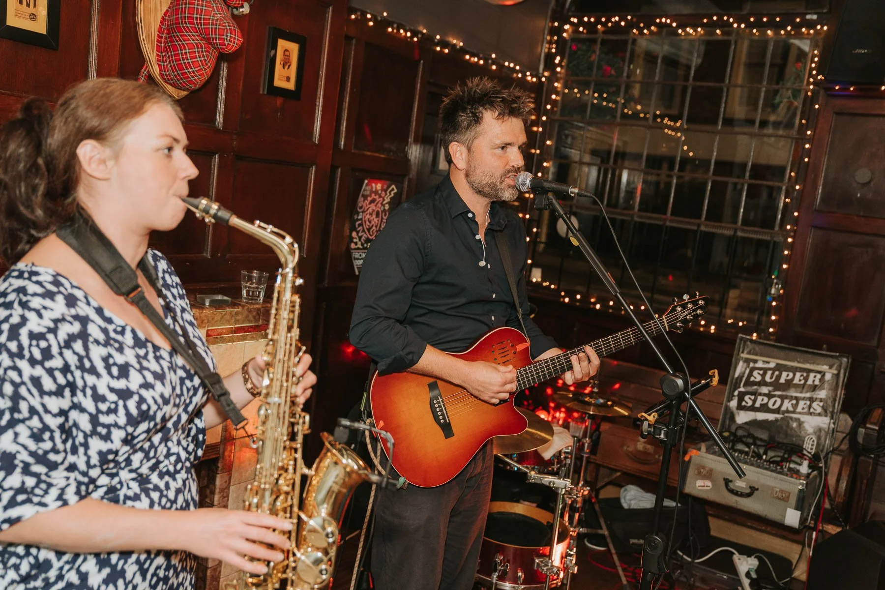  Wedding band performing live at a cosy London pub reception, with a saxophonist and guitarist singing on stage surrounded by warm fairy lights. 