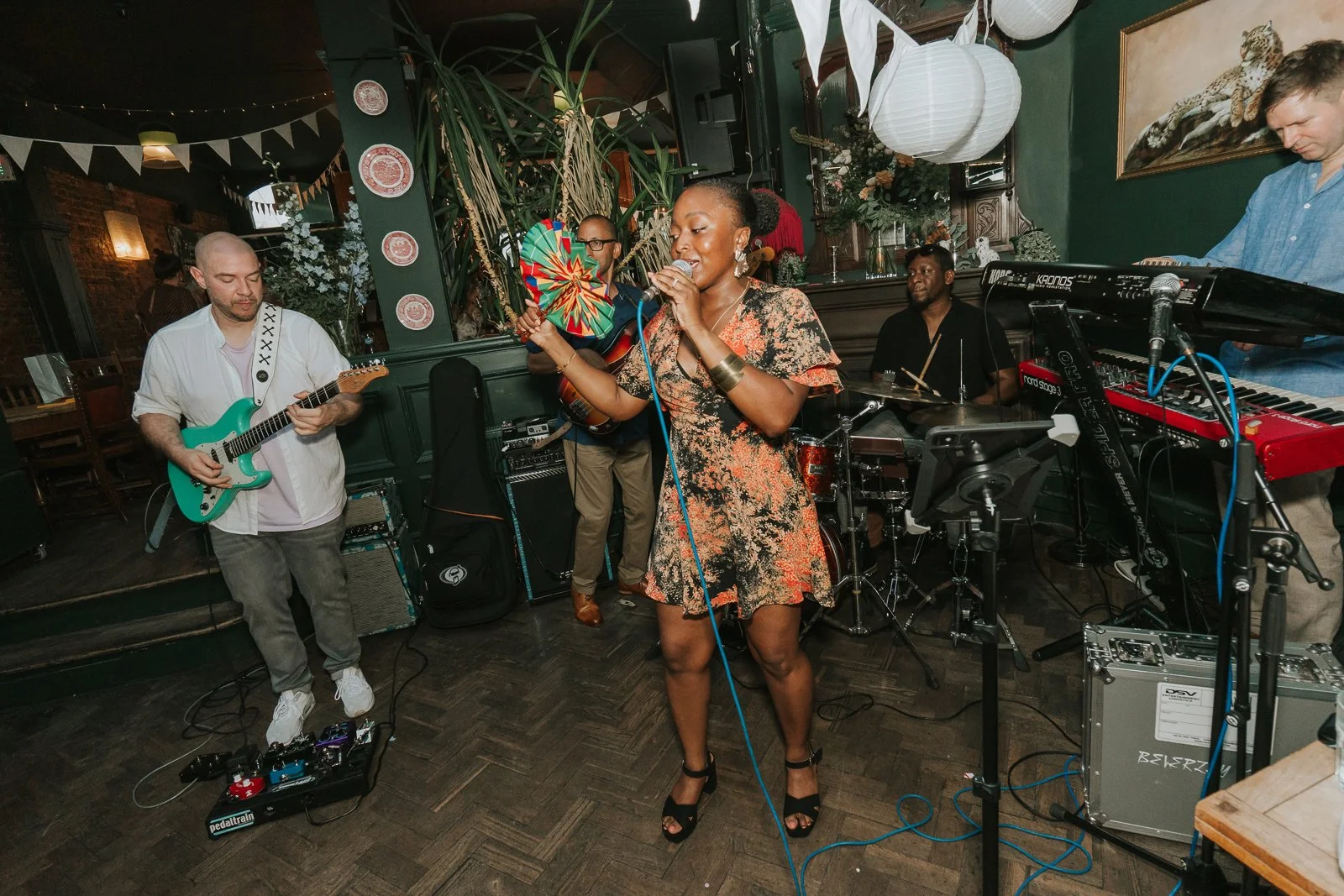  Live wedding band performing at a London pub venue, featuring a singer with a colourful fan and musicians playing guitar, bass and drums. 