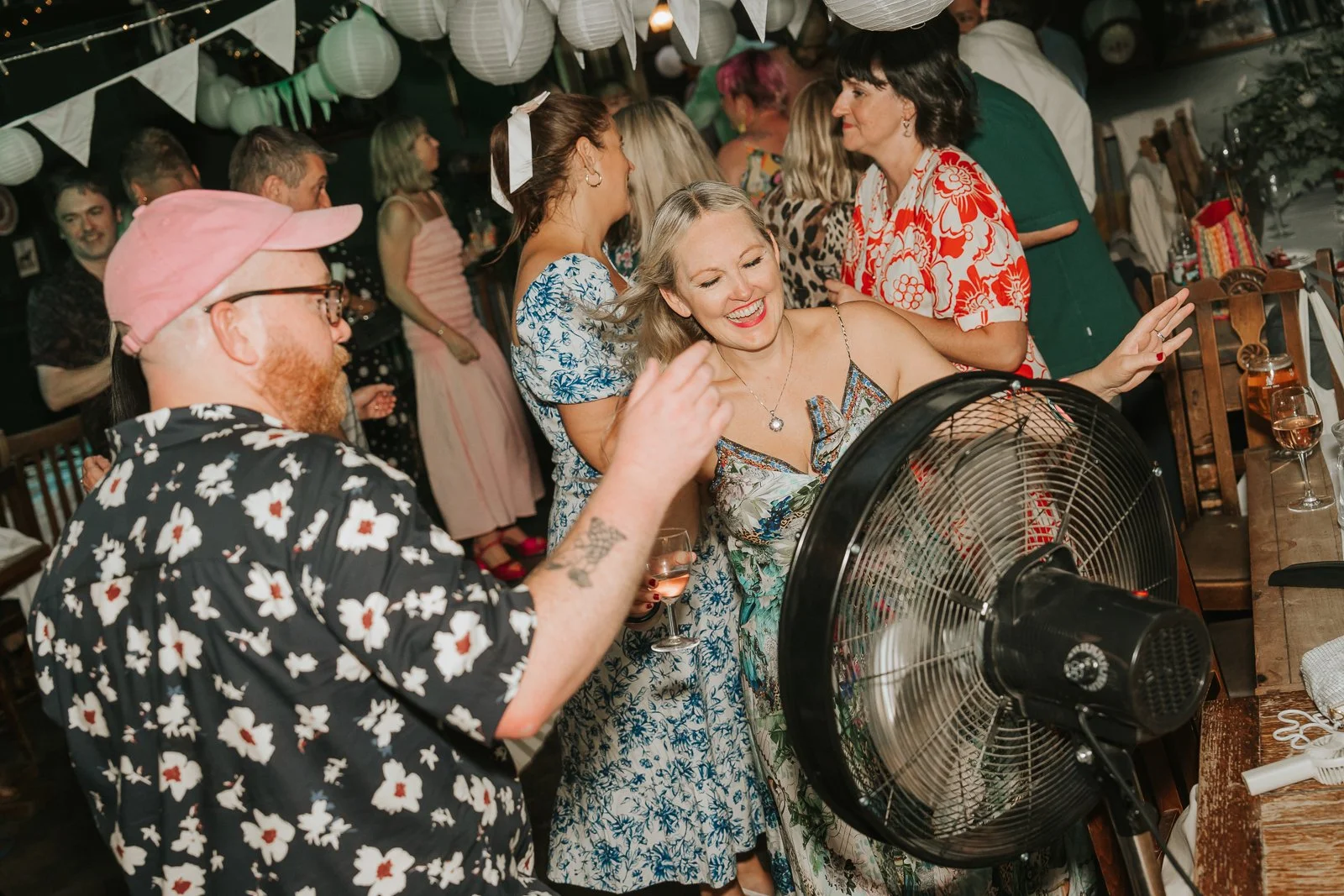  Wedding guests laughing and dancing near a large fan during a busy London pub wedding reception, capturing fun, candid party moments. 