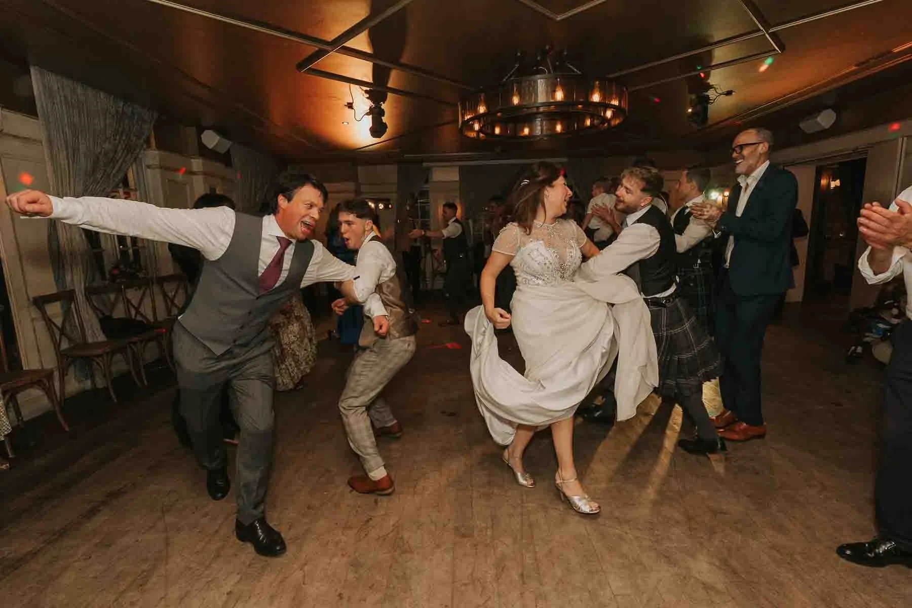  Wedding guests and the bride taking part in energetic ceilidh-style dancing, with joyful movement and laughter on the dancefloor. 