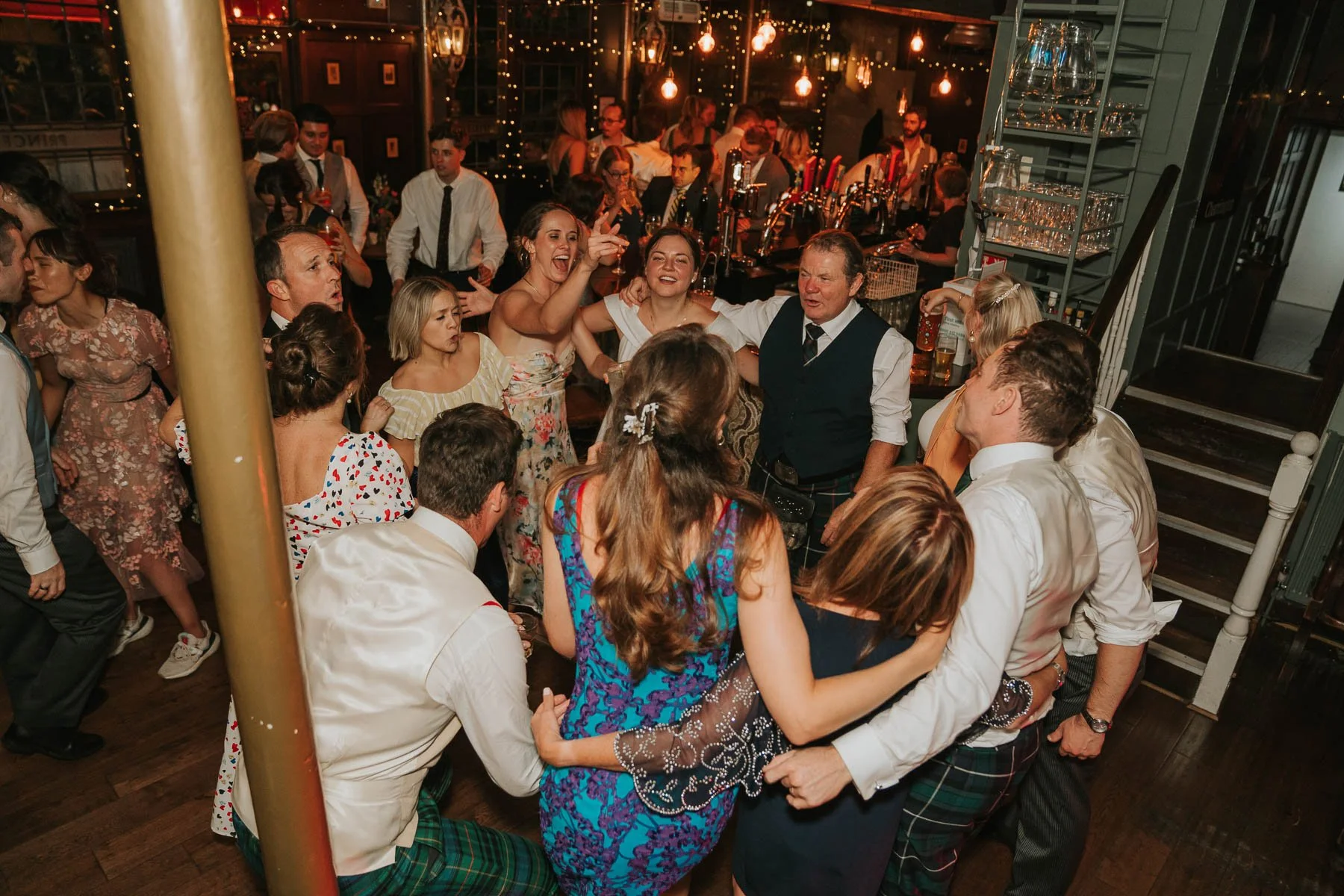  Wedding guests gathered in a lively circle on the dancefloor at a London pub reception, singing, laughing and celebrating together. 