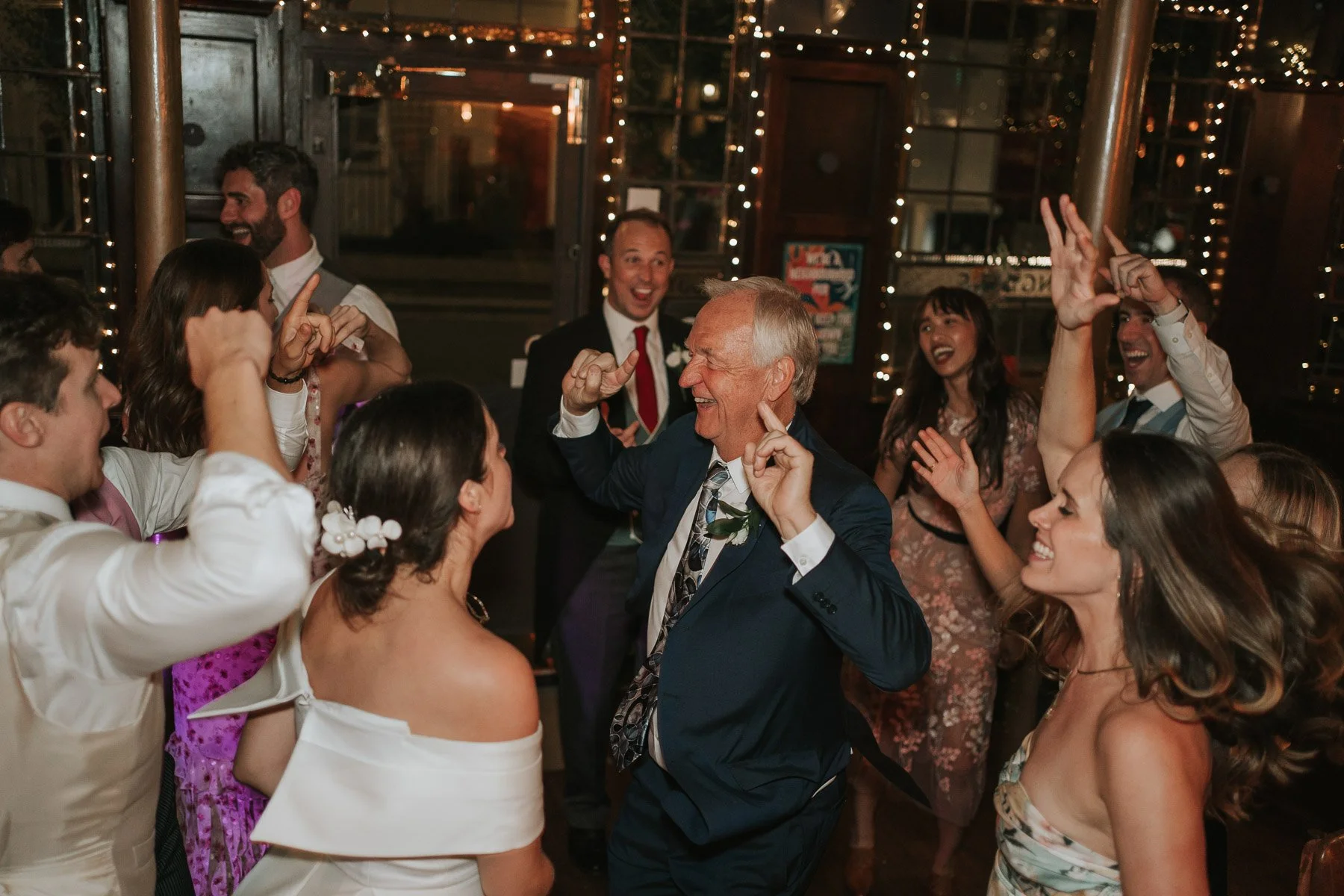  Candid moment of an older guest joyfully dancing with the bride and wedding party during a fun, lively London pub reception. 