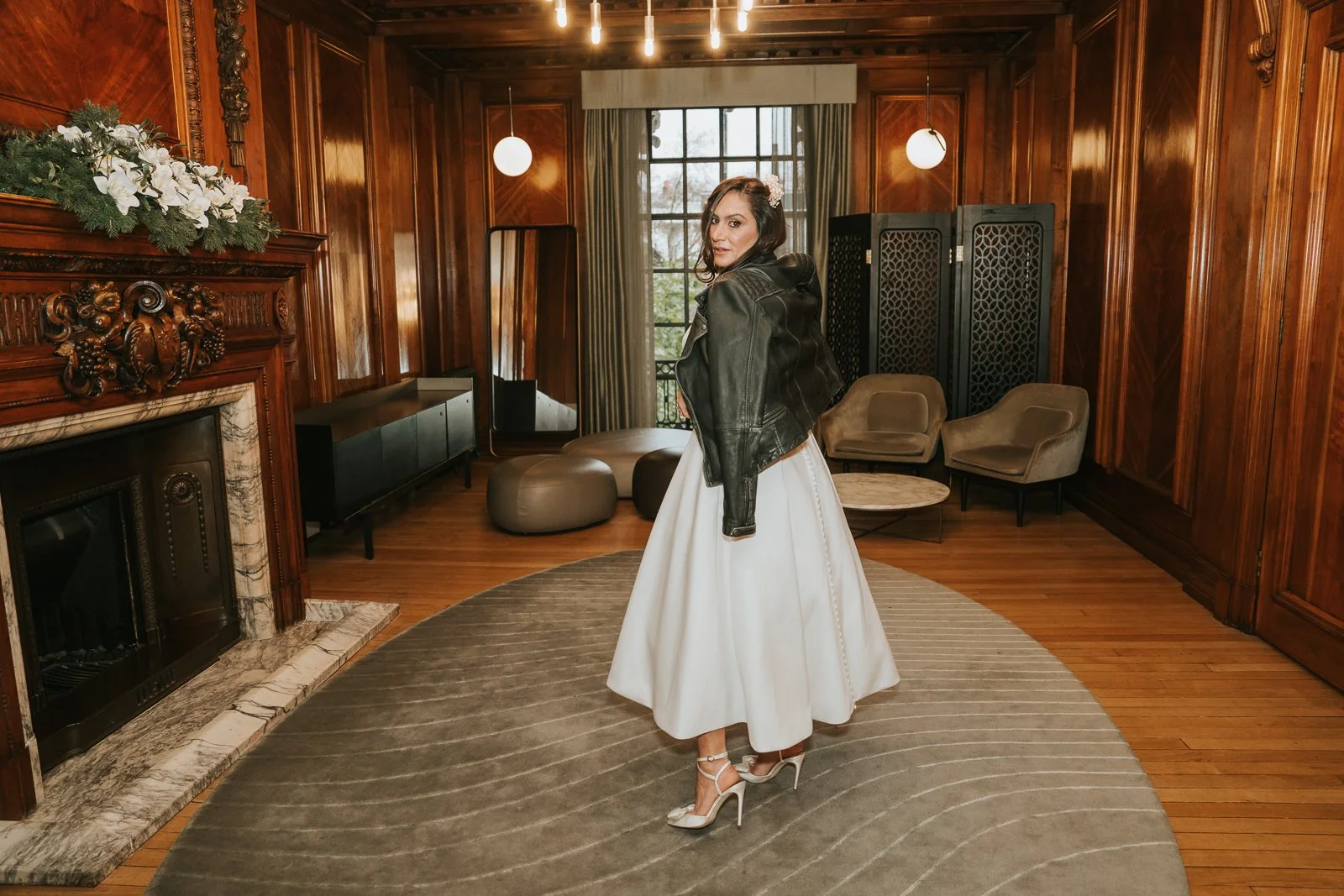  Bride wearing a leather jacket over a modern wedding dress inside Marylebone Town Hall, The Paddington Room, standing in the wood-panelled interior with vintage details and soft natural light. 