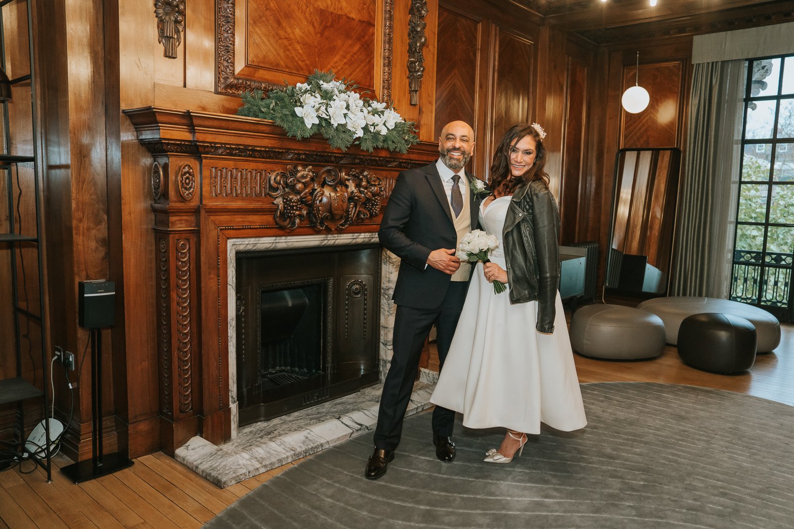  Bride and groom posing together inside Marylebone Town Hall, standing by the ornate fireplace in the elegant wood-panelled Paddington ceremony room. 