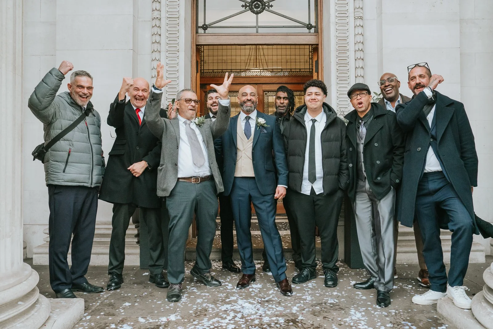  Group of groomsmen celebrating outside Marylebone Town Hall after the wedding ceremony, cheering and laughing on the steps surrounded by confetti. 