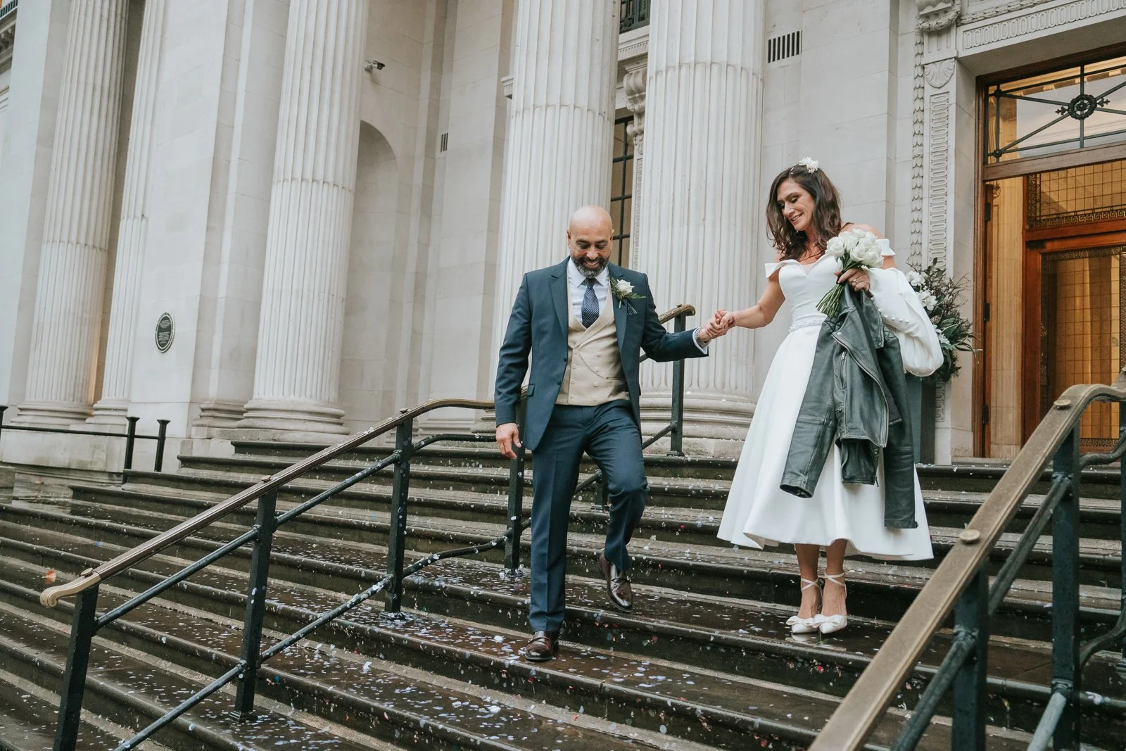  Bride and groom walking hand in hand down the steps of Marylebone Town Hall, confetti scattered around them during their relaxed London wedding exit. 