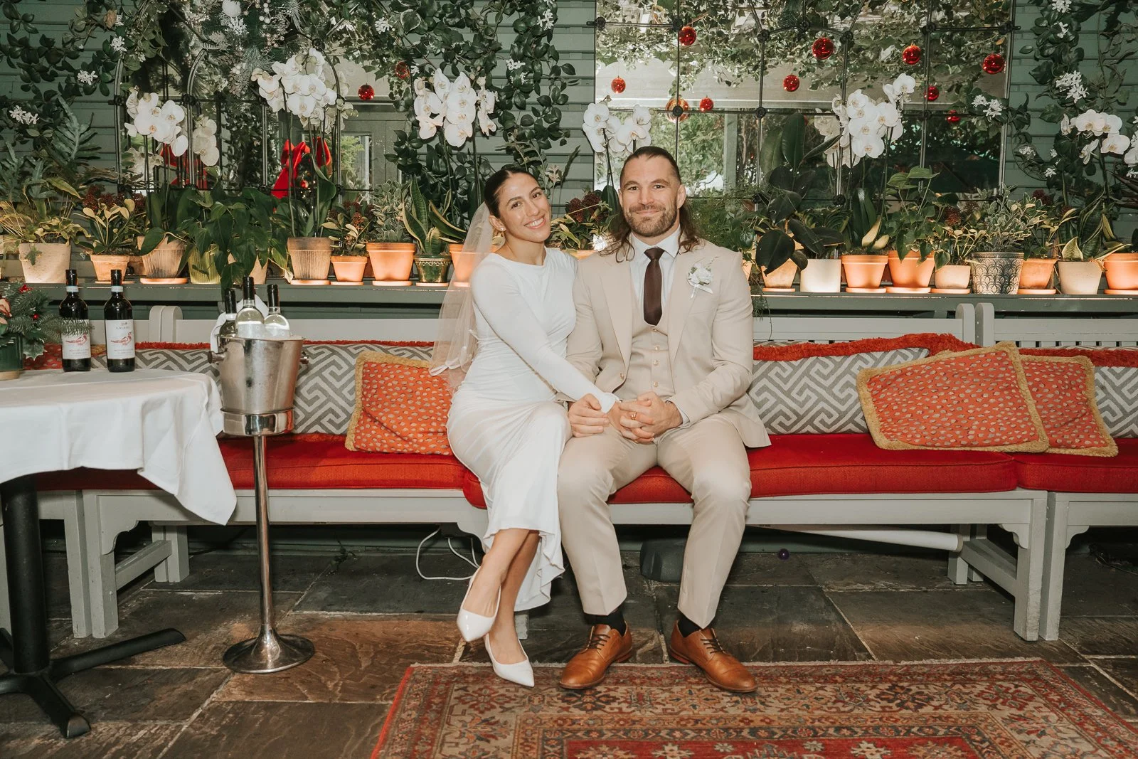  Bride and groom seated together on a bench surrounded by plants and festive décor in the Glasshouse Room at The Ivy Chelsea Garden 